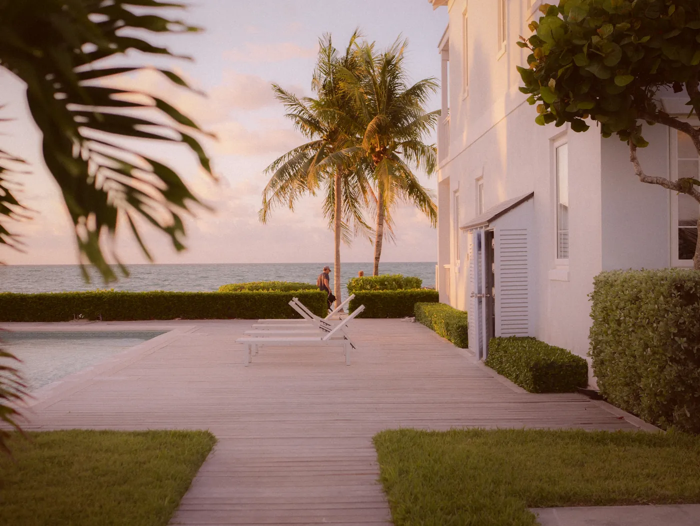 A boardwalk past a white house and palm trees towards the sea at dusk